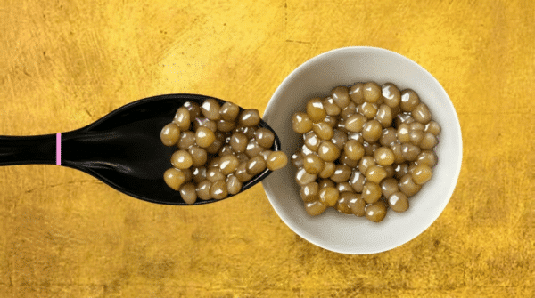 Cuillère noire et coupelle blanche remplies de grains de caviar Schrenki-Dauricus aux reflets dorés sur fond doré.