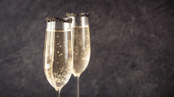 Deux flûtes de champagne pâle, dont les bords sont décorés de grains de caviar, devant un fond sombre.