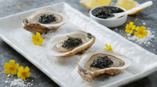 Trois huîtres ouvertes sur glace garnies de caviar, avec une coupelle de caviar en arrière-plan.